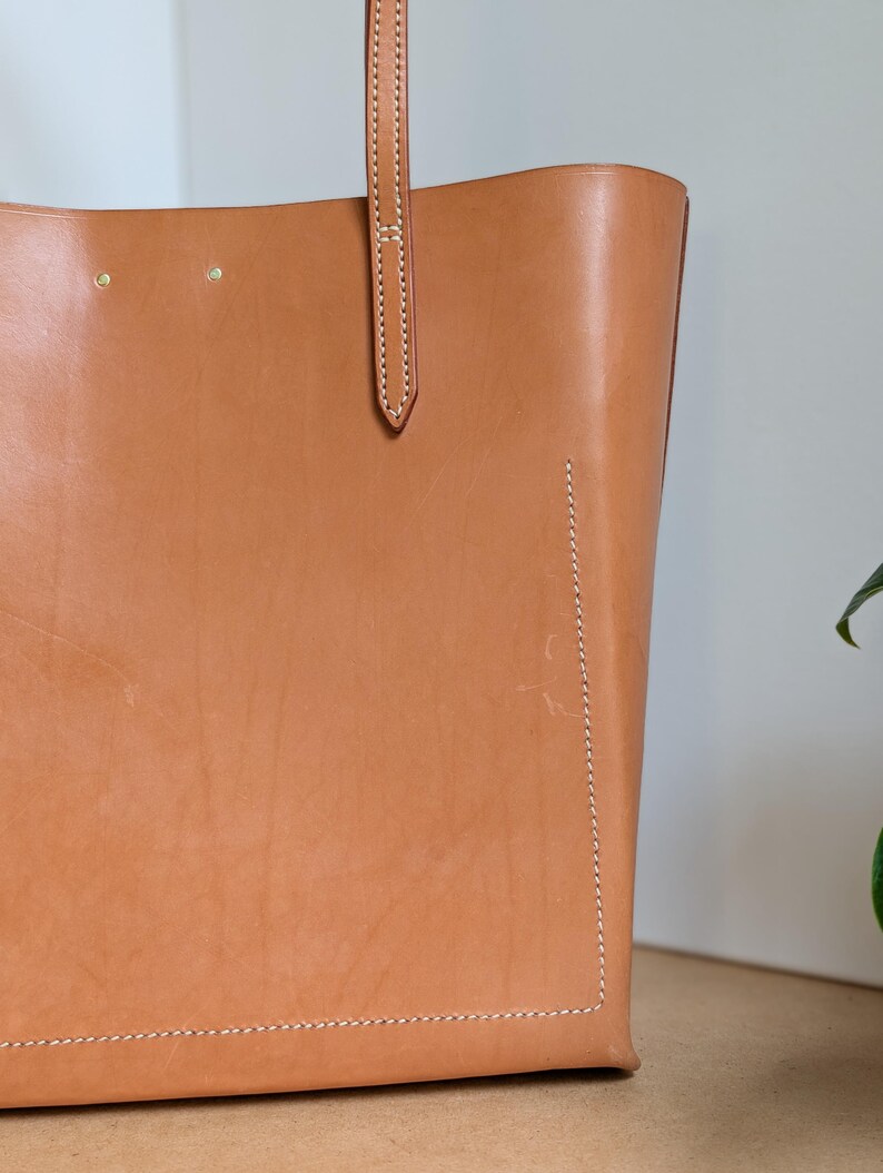 May include: A tan leather tote bag featuring visible white stitching along the edges and a vertical strap attachment. The surface displays natural grain textures and two small metal rivets near the top edge of the bag's main body.