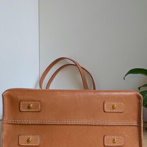 May include: A tan leather handbag featuring a rectangular base with four brass-toned metal feet, each secured by a rectangular leather patch with white contrast stitching. Two thin leather handles extend upward from the top of the bag.