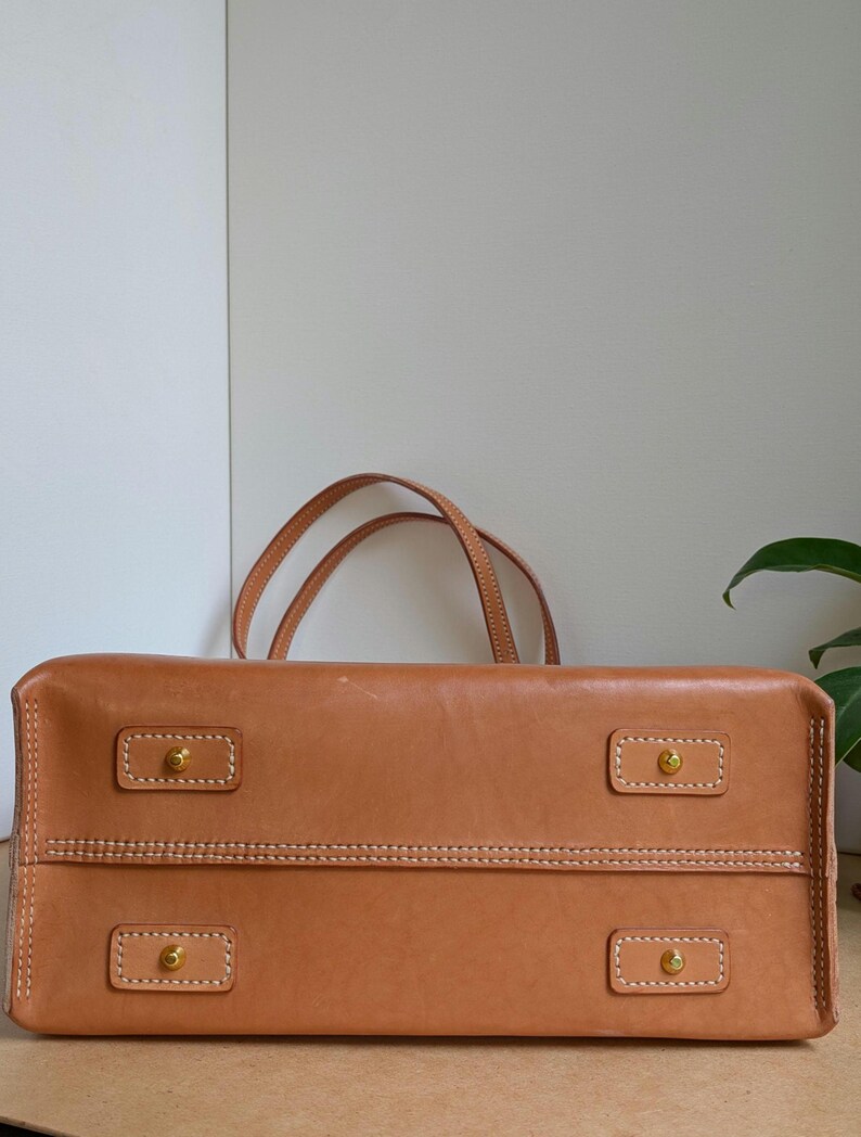 May include: A tan leather handbag featuring a rectangular base with four brass-toned metal feet, each secured by a rectangular leather patch with white contrast stitching. Two thin leather handles extend upward from the top of the bag.