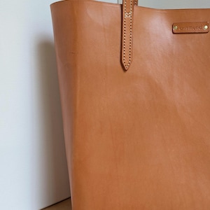 May include: A tan leather tote bag featuring vertical stitching on the handle attachment and a rectangular leather patch embossed with the text ansmund secured by two small brass-colored rivets on the upper right side of the bag exterior.
