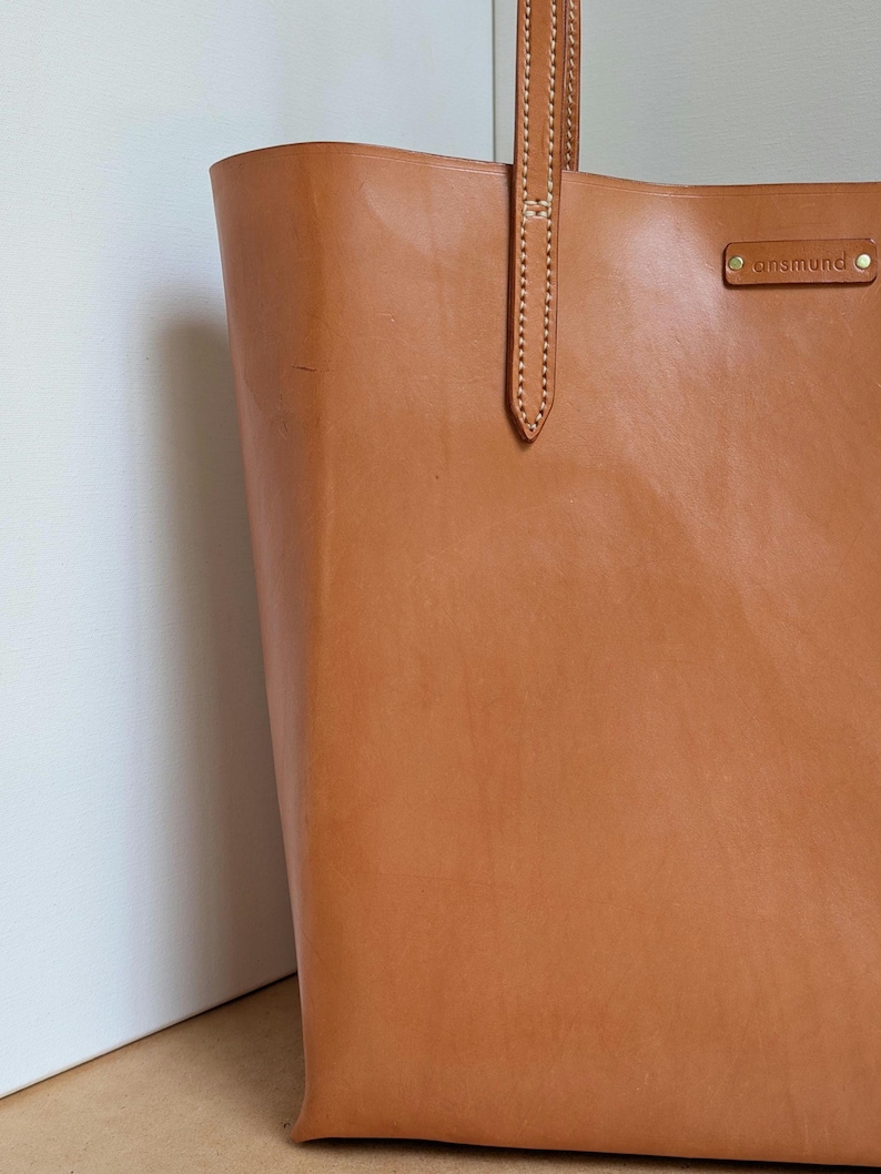 May include: A tan leather tote bag featuring vertical stitching on the handle attachment and a rectangular leather patch embossed with the text ansmund secured by two small brass-colored rivets on the upper right side of the bag exterior.