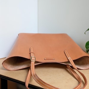 May include: A tan leather tote bag featuring two long shoulder straps with brass-toned metal buckles and white contrast stitching. The bag has an open top design with a smooth, unlined interior and a small rectangular leather brand tag inside.