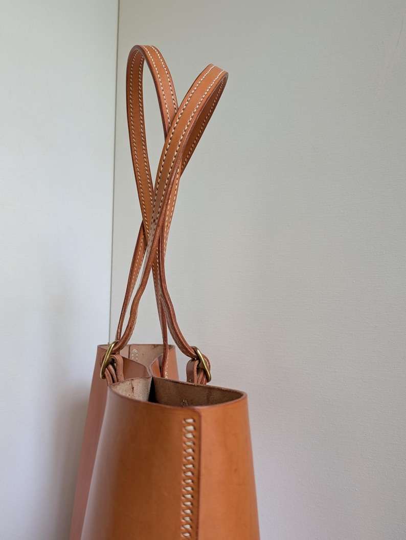 May include: A tan leather bucket bag featuring two long, slender handles attached with brass-toned metal hardware. The bag displays visible white stitching along the edges and the handle seams, with a smooth, structured leather construction.
