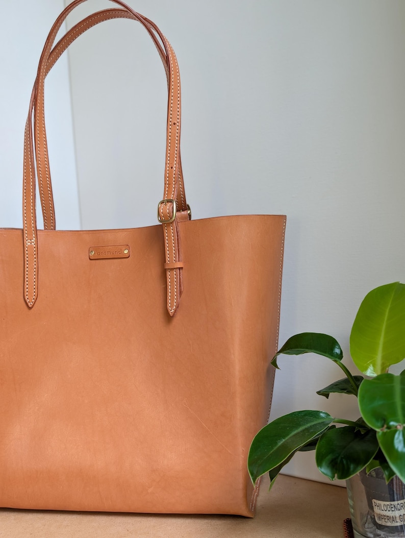 May include: A tan leather tote bag featuring two long shoulder straps with adjustable gold-tone metal buckles. The front of the bag displays a small rectangular leather patch embossed with the text "antmund" and secured with two gold-tone rivets.
