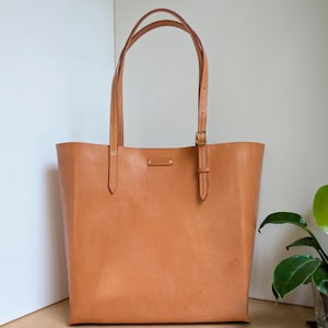 May include: A tan leather tote bag with two thin shoulder straps featuring adjustable metal buckles. The bag has a rectangular shape with visible stitching along the handles and a small rectangular brand tag centered near the top opening. Text reads Bag No.2.