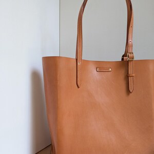 May include: A tan leather tote bag featuring two long shoulder straps with adjustable brass buckles, visible white stitching along the edges, and a small rectangular metal brand tag centered near the top opening of the bag.