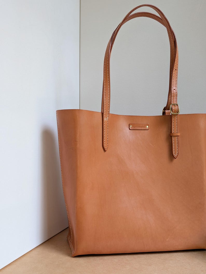 May include: A tan leather tote bag featuring two long shoulder straps with adjustable brass buckles, visible white stitching along the edges, and a small rectangular metal brand tag centered near the top opening of the bag.