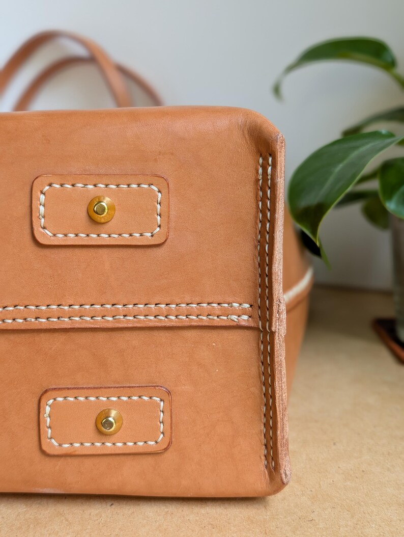 May include: A tan leather bag base featuring two rectangular leather patches, each secured with a central brass-toned metal stud. The edges are reinforced with thick, visible white stitching along the seams and the perimeter of the patches.