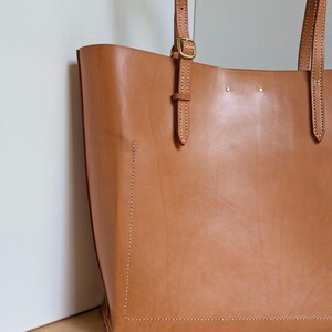 May include: A tan leather tote bag featuring visible white stitching along the edges, two adjustable shoulder straps with brass-toned metal buckles, and two small brass-toned rivets near the top center of the bag's front panel.