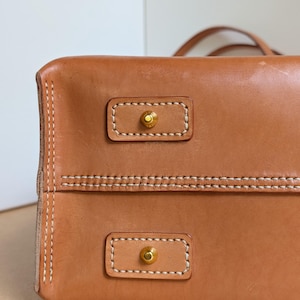 May include: A tan leather bag base featuring two rectangular leather patches, each secured with white contrast stitching and centered with a small, circular gold-toned metal stud. The leather surface shows a smooth texture with visible stitching.