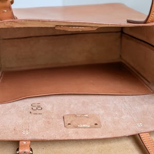 May include: A tan leather tote bag with an open top, featuring visible stitching, two leather handles, and a smooth interior pocket. The interior base shows a stamped numerical code 031 on the light-colored, unfinished leather surface.