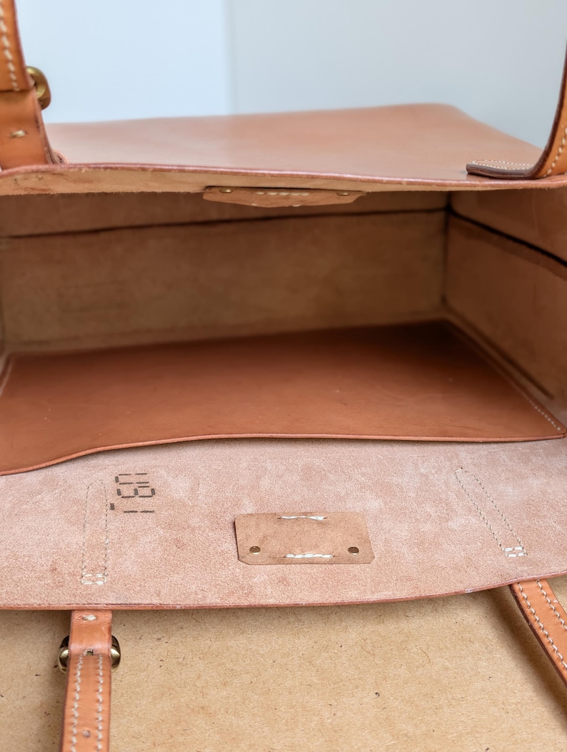 May include: A tan leather tote bag with an open top, featuring visible stitching, two leather handles, and a smooth interior pocket. The interior base shows a stamped numerical code 031 on the light-colored, unfinished leather surface.