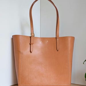 May include: A tan leather tote bag featuring two long shoulder straps with adjustable brass buckles, visible white contrast stitching along the bottom and sides, and two small brass rivets at the top center of the front panel.
