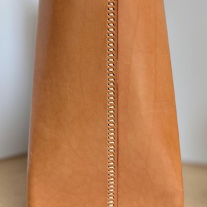 May include: A tan leather object featuring a vertical seam with visible, light-colored stitching running down the center. The leather has a smooth, natural texture with subtle variations in tone, and the base is finished with a horizontal stitched edge.