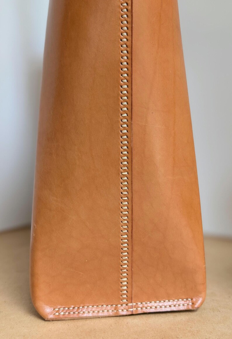 May include: A tan leather object featuring a vertical seam with visible, light-colored stitching running down the center. The leather has a smooth, natural texture with subtle variations in tone, and the base is finished with a horizontal stitched edge.
