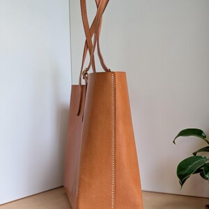 May include: A tan leather tote bag with two tall, thin shoulder straps. The bag features visible light-colored stitching along the side seams and the strap attachments. The structure is upright and rectangular with a smooth, matte finish.