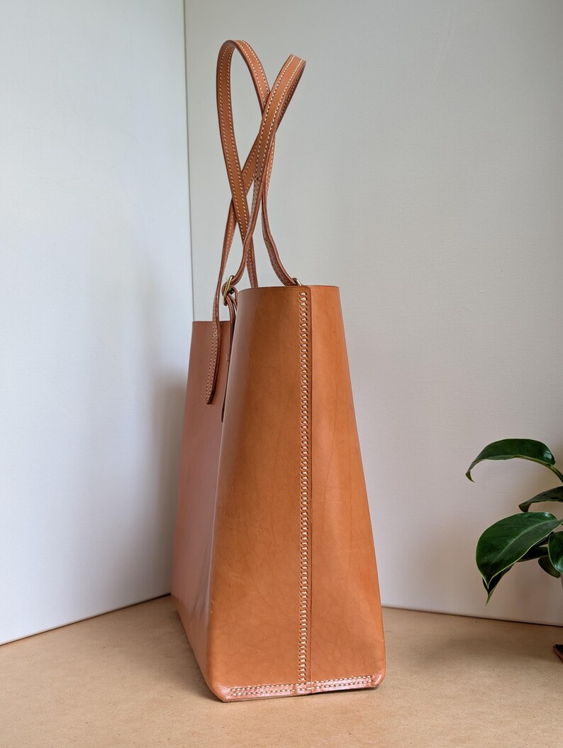 May include: A tan leather tote bag with two tall, thin shoulder straps. The bag features visible light-colored stitching along the side seams and the strap attachments. The structure is upright and rectangular with a smooth, matte finish.