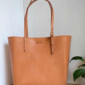 May include: A tan leather tote bag featuring two long, adjustable shoulder straps with brass-colored buckles and a small rectangular leather brand tag centered near the top opening. The bag has visible white stitching along the straps and body.