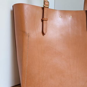 May include: A tan leather tote bag featuring visible light-colored stitching along the bottom and side panels. The bag includes thin leather shoulder straps with adjustable brass-toned metal buckles and two small brass rivets near the top edge.