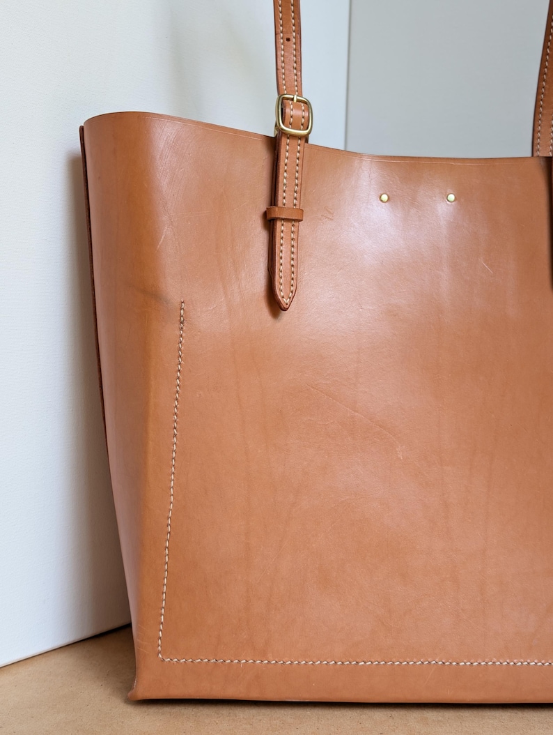 May include: A tan leather tote bag featuring visible light-colored stitching along the bottom and side panels. The bag includes thin leather shoulder straps with adjustable brass-toned metal buckles and two small brass rivets near the top edge.