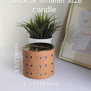 May include: A tan candle holder with a gold-colored lid. The holder has a cylindrical shape with evenly spaced circular cutouts. It measures 3 1/2 inches (9cm) tall and 4 1/4 inches (10.5cm) wide. The text on the image says "Fits B&BW 3wick or smaller size candle."