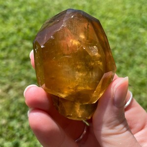 May include: A faceted, golden-brown crystal held against a backdrop of green grass. The crystal has a translucent quality, with visible internal structures and a polished appearance. The light catches the facets, creating highlights and shadows.