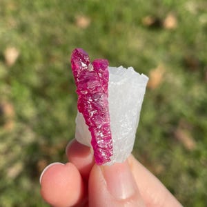 May include: A raw mineral specimen featuring a vibrant, deep magenta-colored crystal formation attached to a jagged, translucent white crystalline base. The magenta crystal has a textured, multifaceted surface with a distinct vertical orientation.