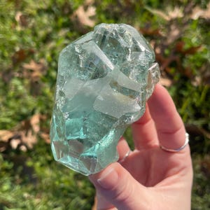 May include: A large, irregularly shaped, translucent aquamarine crystal. The crystal has multiple facets and a slightly rough texture. The background is blurred, showing green grass and fallen leaves.
