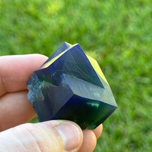 May include: A dark blue and green crystal with multiple facets. The crystal is shaped like a pyramid and has a smooth, polished surface.
