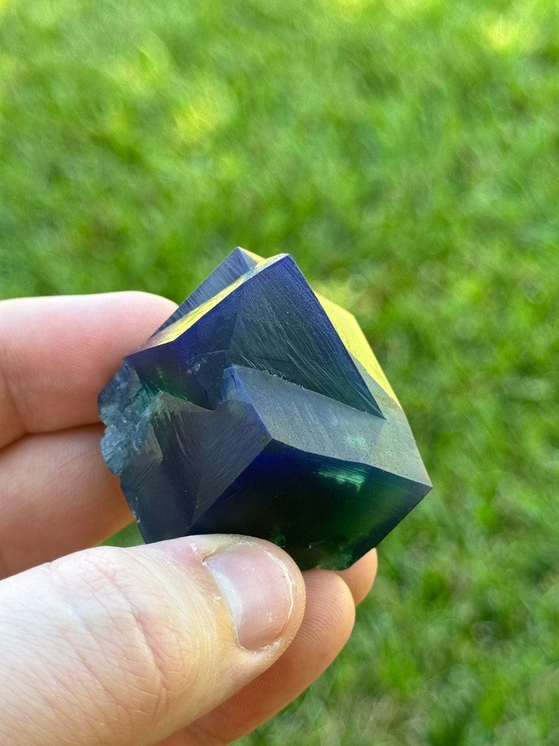 May include: A dark blue and green crystal with multiple facets. The crystal is shaped like a pyramid and has a smooth, polished surface.