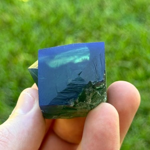 May include: A dark green and blue fluorite crystal with a cube shape. The crystal is translucent and has a slight shimmer.