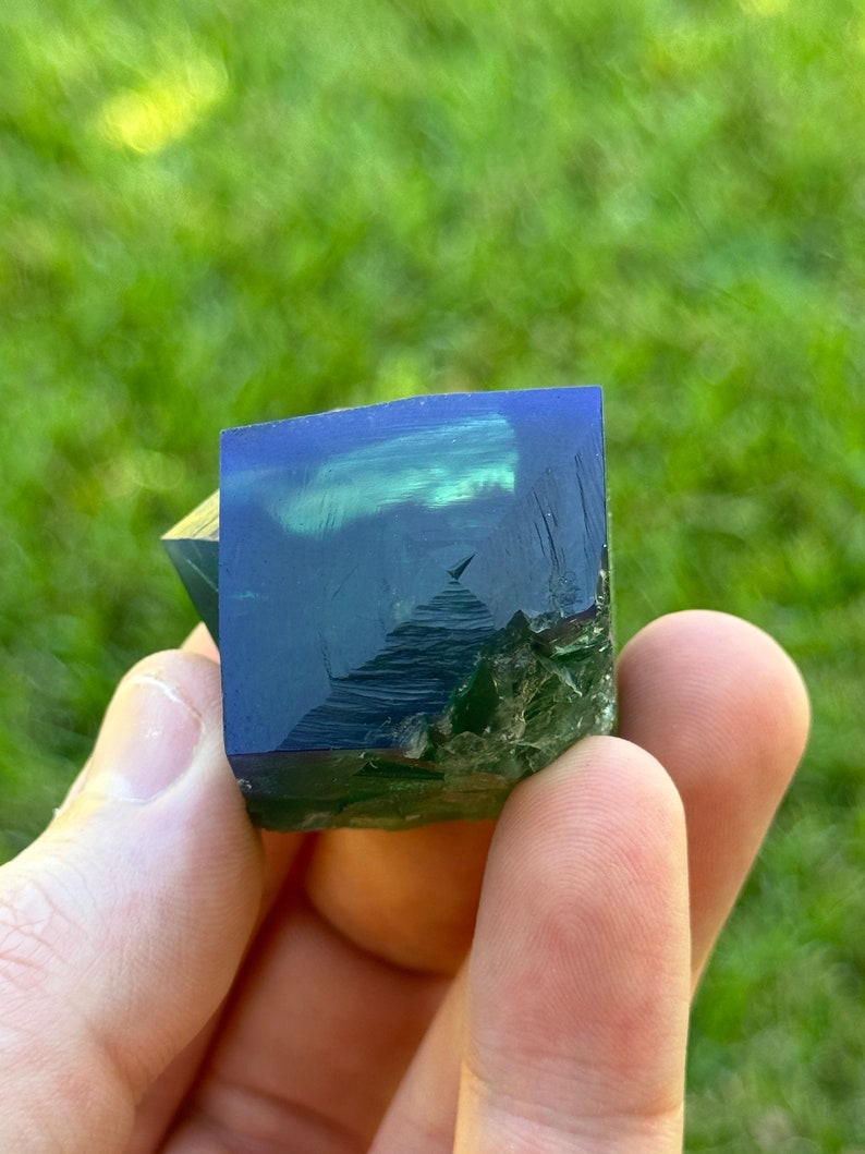 May include: A dark green and blue fluorite crystal with a cube shape. The crystal is translucent and has a slight shimmer.