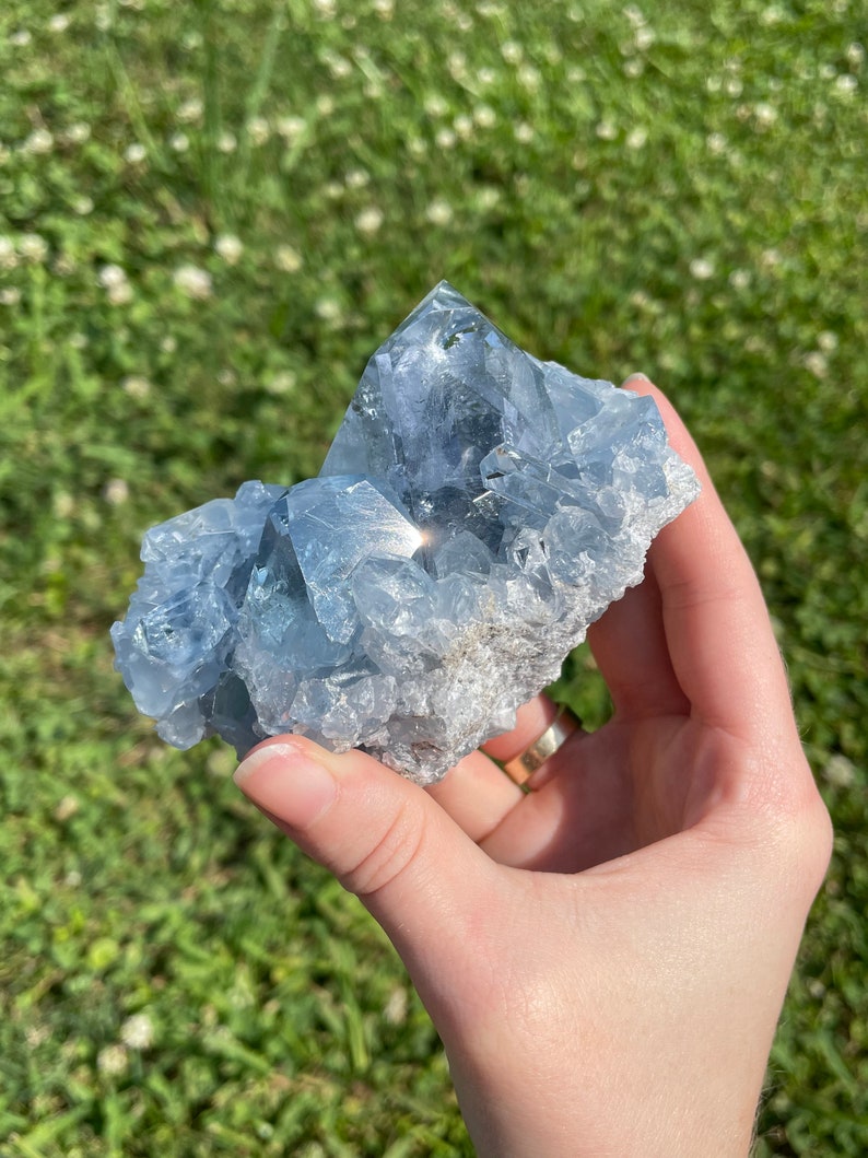 May include: A large, light blue celestite crystal cluster with a crystalline structure. The crystal has a rough, textured base and is held against a background of green grass.