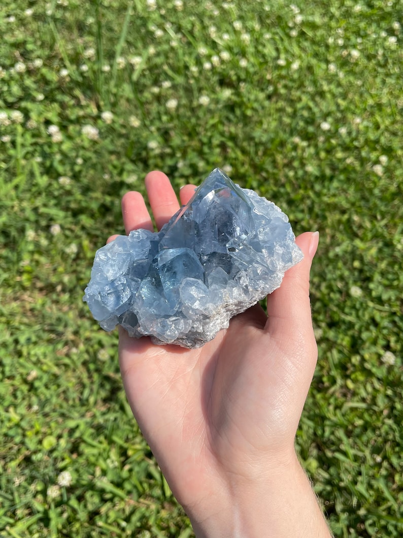 May include: A large, light blue celestite crystal cluster. The crystal has a rough, textured surface and a crystalline structure. The cluster is held in a hand against a background of green grass.
