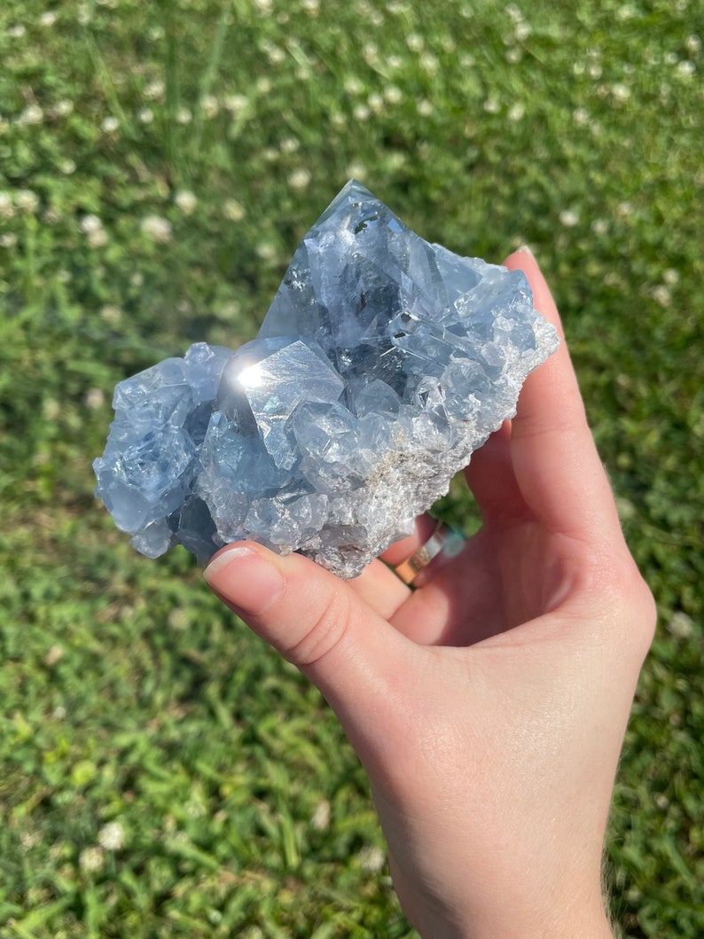 May include: A light blue celestite crystal cluster. The crystal has a crystalline structure with a rough, textured surface. The color is a pale, translucent blue with areas of white. The crystal is held against a background of green grass.