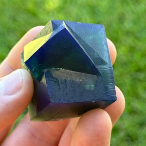 May include: A dark blue and green geometric crystal with a faceted design. The crystal is held in a hand.