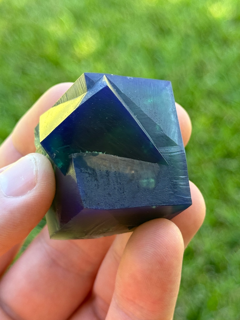 May include: A dark blue and green geometric crystal with a faceted design. The crystal is held in a hand.