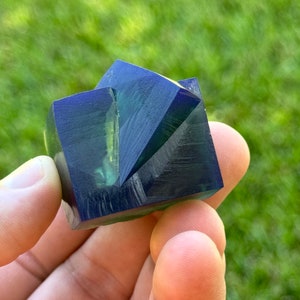 May include: A dark blue and green crystal with a geometric shape. The crystal has a smooth, polished surface and is faceted.