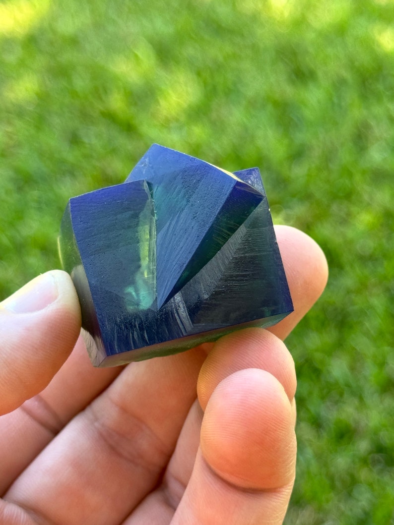 May include: A dark blue and green crystal with a geometric shape. The crystal has a smooth, polished surface and is faceted.