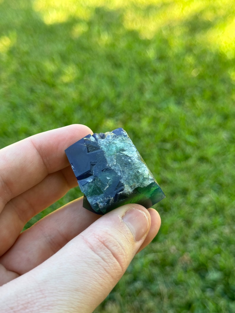 May include: A dark blue and green fluorite crystal with a rough, natural texture. The crystal is a geometric shape with many facets.