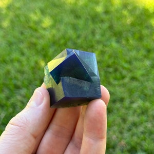 May include: A deep blue and green fluorite crystal with multiple facets. The crystal is a cube shape and is being held in a hand.