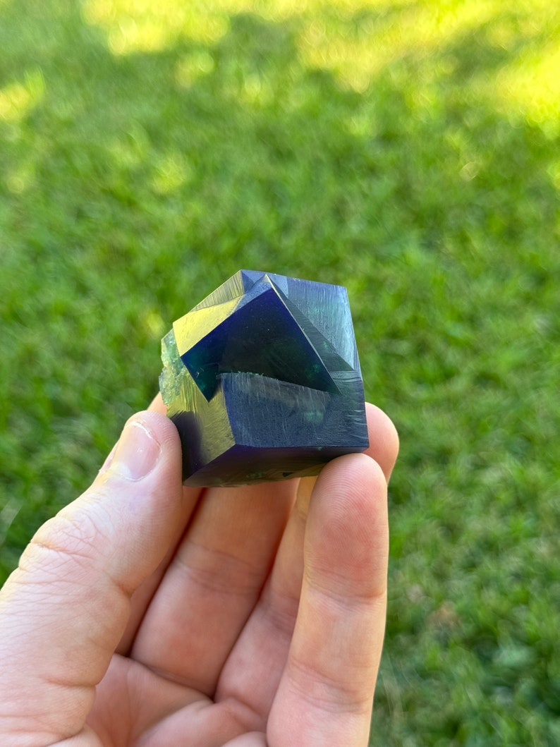 May include: A deep blue and green fluorite crystal with multiple facets. The crystal is a cube shape and is being held in a hand.