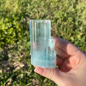 May include: A light blue aquamarine crystal with a rectangular shape and vertical striations. The crystal is held in a hand against a blurred green background. The crystal's color is a pale, translucent blue.
