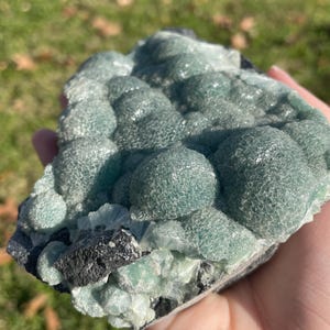 May include: A raw mineral specimen featuring clusters of rounded, translucent, sea-green botryoidal formations. The surface of these spheres displays a fine, crystalline texture, contrasted by dark, jagged, charcoal-coloured rock at the base.