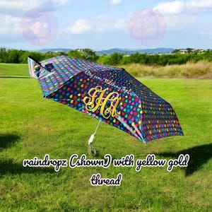 May include: A black umbrella with a colorful raindrop pattern and a gold monogram. The umbrella is open and the handle is visible. The text "Etsy.com/StitchingTheLove" is printed on the umbrella. The umbrella is on a green grassy field.