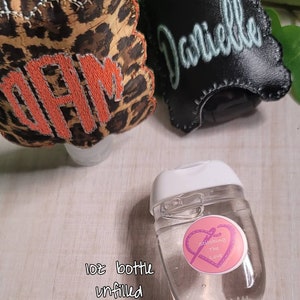 Może przedstawiać: Przezroczysta plastikowa butelka o pojemności 30 ml z białą zakrętką i r&oacute;żowo-fioletową naklejką z napisem "Stitching the Love". Butelka jest pusta i stoi na drewnianej powierzchni. W tle znajdują się dwa przedmioty wykonane z tkaniny, jeden z nadrukiem lamparta i wyszytymi literami "DAM", a drugi jest czarny z wyszytym imieniem "Danielle".