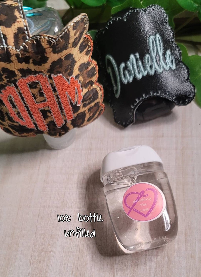 Może przedstawiać: Przezroczysta plastikowa butelka o pojemności 30 ml z białą zakrętką i r&oacute;żowo-fioletową naklejką z napisem "Stitching the Love". Butelka jest pusta i stoi na drewnianej powierzchni. W tle znajdują się dwa przedmioty wykonane z tkaniny, jeden z nadrukiem lamparta i wyszytymi literami "DAM", a drugi jest czarny z wyszytym imieniem "Danielle".