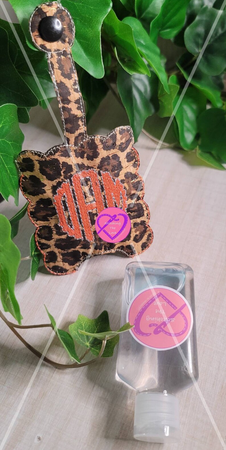 Może przedstawiać: Brelok do kluczy w panterkę z monogramem "OPM" wyszytym pomarańczową nicią. Brelok ma czarny guzik na g&oacute;rze. Obok breloka znajduje się mała, przezroczysta butelka z płynem do dezynfekcji rąk z r&oacute;żową etykietą z napisem "Stitching the Love".