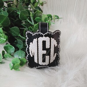 Może przedstawiać: Czarny sk&oacute;rzany brelok do kluczy z haftowanym białym monogramem "MEC" w k&oacute;łku z falistą obw&oacute;dką. Tekst "Stitchingthelove" jest wyszyty pod monogramem.