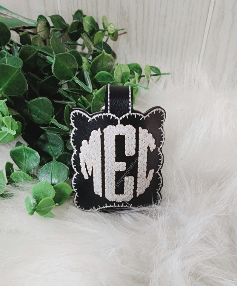 Może przedstawiać: Czarny sk&oacute;rzany brelok do kluczy z haftowanym białym monogramem "MEC" w k&oacute;łku z falistą obw&oacute;dką. Tekst "Stitchingthelove" jest wyszyty pod monogramem.